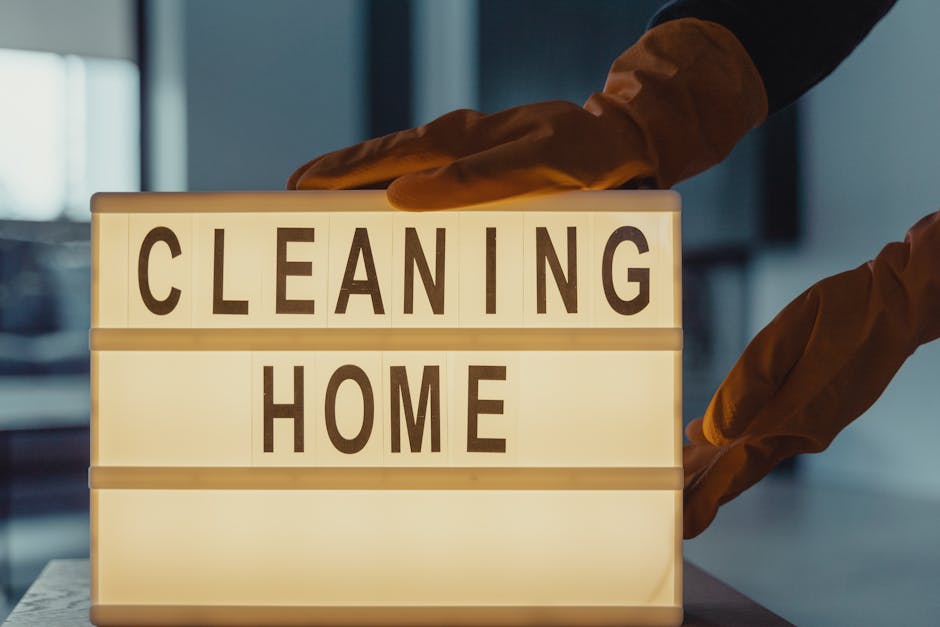 A person wearing brown cleaning gloves is placing a lit marquee sign that displays the words 'CLEANING HOME' in black letters. The sign is illuminated from within, creating a soft glow against the bac
