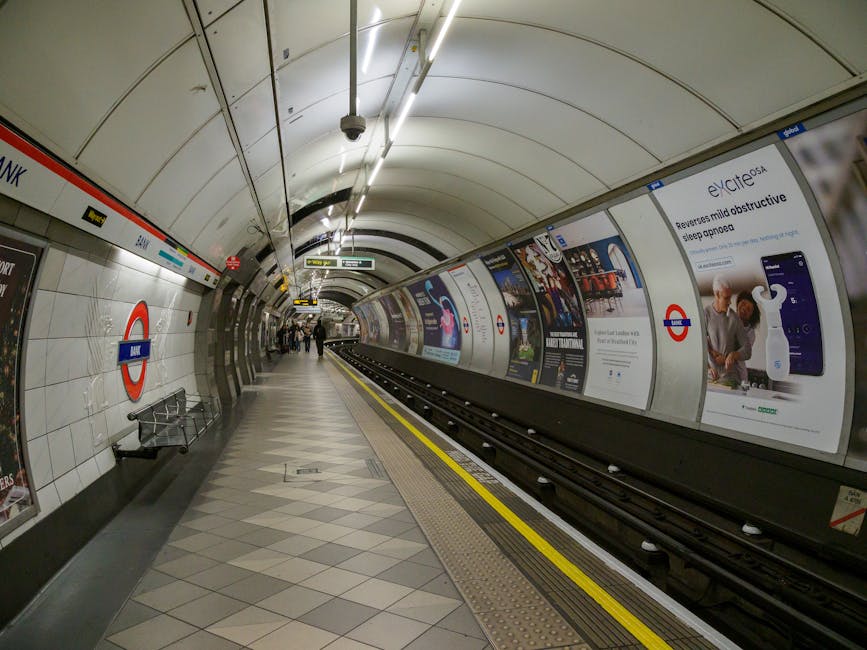 An underground station platform at High Barnet tube station with clean, tiled floors and smooth, curved white walls. The platform features a row of advertisements displayed along the wall, some showca