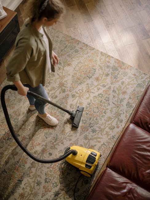 A professional cleaning technician dressed in white protective coveralls, a face mask, and blue gloves is performing surface cleaning in a residential living room. The technician is using a machine th
